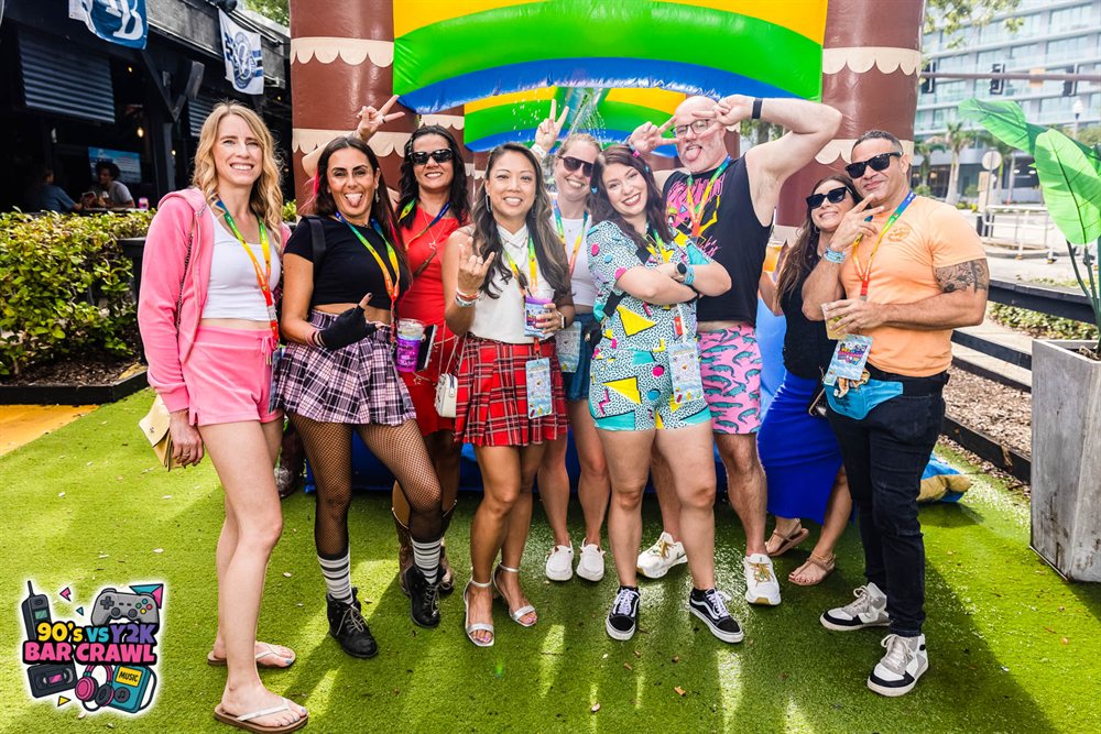 Nine friends in 90s outfits posing under the inflatable palm-tree arch at the 90s vs Y2K Bar Crawl